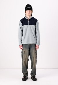 Pullover grigio in fleece con pannello superiore blu navy, collo a zip, abbinato a pantaloni cargo larghi e a un berretto nero con testo bianco.