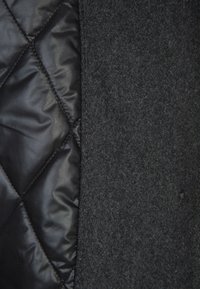 Gros plan sur deux textures de tissu : matériau noir brillant matelassé à gauche et laine douce gris foncé à droite, reliés par une couture.