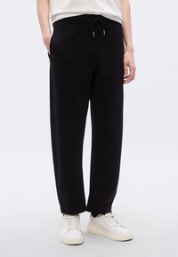 Μαύρο παντελόνι φόρμας με ελαστική μέση και κορδόνι, με πλαϊνές τσέπες και άνετη εφαρμογή, συνδυασμένο με λευκά αθλητικά παπούτσια.