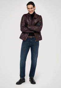 Chaqueta de cuero marrón sobre un cuello de tortuga negro, combinada con jeans azules y botas de tobillo negras. Modelo de pie con los brazos cruzados, sonriendo.