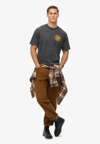 Szary t-shirt z krótkim rękawem z żółtą grafiką, brązowe spodnie z kordurowej tkaniny i koszula w kratę przewiązana w talii. Czarne buty i zegarek.