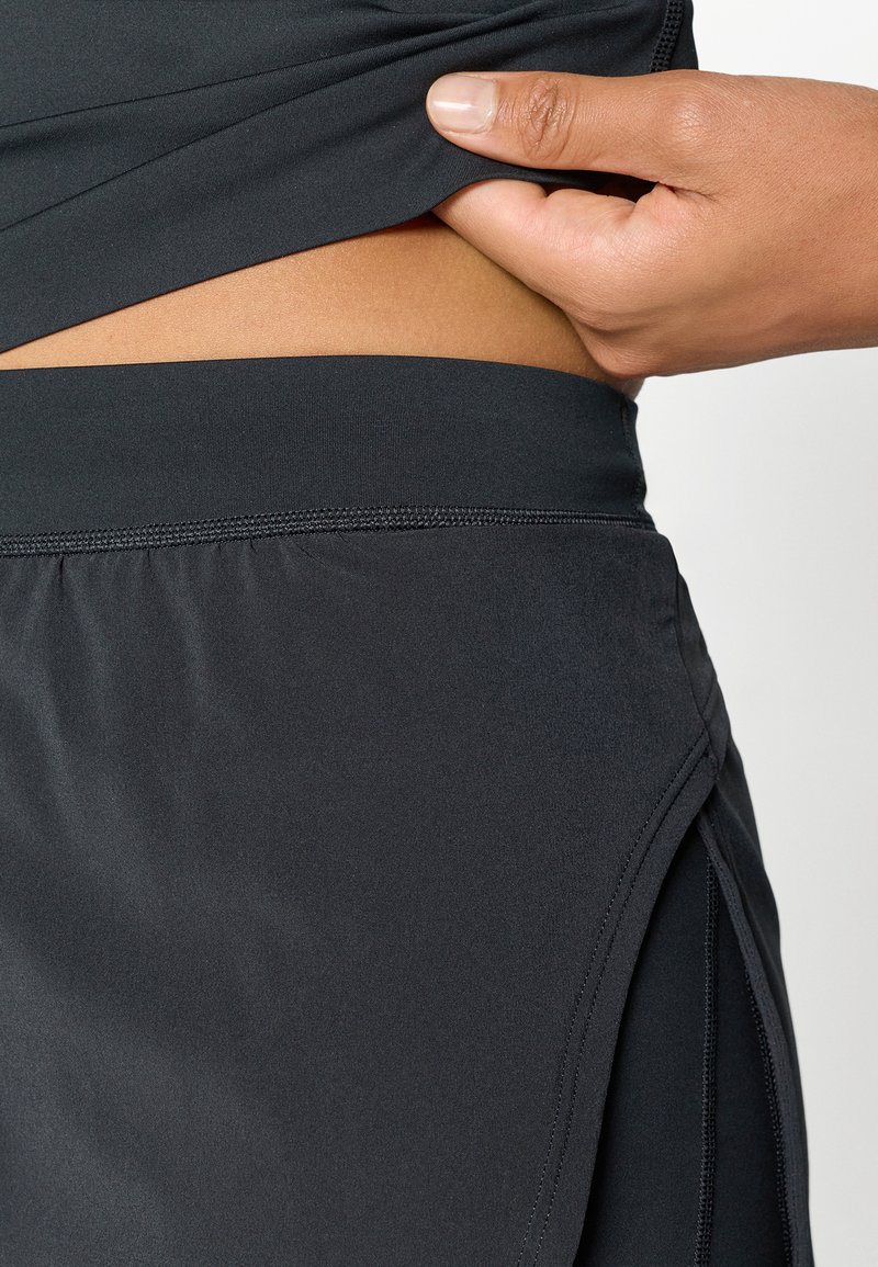 Black athletic top with a smooth texture, featuring a side slit and an elastic waistband. Hand lifting the fabric for visibility.