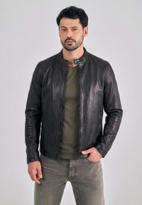 Mężczyzna z ciemnymi włosami i brodą, ubrany w czarn ą skórzaną kurtkę, oliwkowozielony t-shirt i wyblakłe szare dżinsy, stojący z jedną ręką w kieszeni.