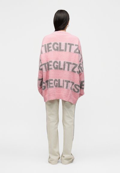 Personne aux longs cheveux foncés portant un pull rose oversize avec un texte gris en gras et un pantalon écru, debout de dos sur fond blanc.