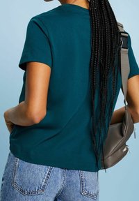 Teal kortärmad bomullströja med rund halsringning och avslappnad passform, tillsammans med ljusblå denimjeans och en grå axelväska.