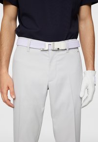 Nämä on värjätyt valkoisesta nahasta valmistettu punottu vyö, jossa on valkoinen metallinen solki. Vyö on yhdistetty vaaleanharmaisiin housuihin ja valkoiseen käsineeseen. Taustalla näkyy tumma paita.