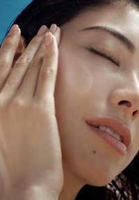 Primo piano del volto di una donna con pelle liscia, labbra leggermente tinte e lunghi capelli scuri. Tiene delicatamente la guancia con una mano, gli occhi chiusi.