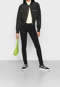 Chaqueta bomber negra con bolsillos con cremallera, suéter de cuello alto color crema, jeans negros ajustados y zapatillas negras. Lleva un pequeño bolso verde.