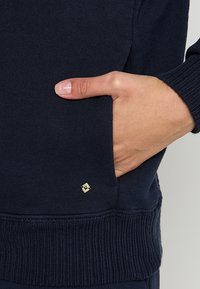 Sweat-shirt bleu marine en tissu doux, avec des poignets et un ourlet côtelés, et une étiquette de logo dorée près de la poche avant.