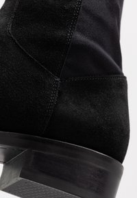 Botín negro de ante y suave tejido, con un tacón apilado y una textura lisa que presenta un cuidado detalle de costura.