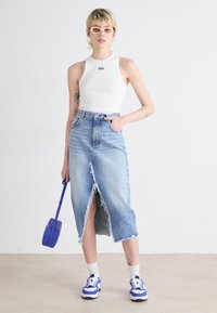 Vit ärmlös topp, högmidjad jeanskjol med fransad nederkant och framspricka, blå sneakers, håller en blå accessoar.
