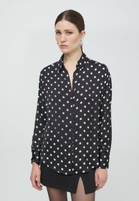 Camicia nera con bottoni, con pois bianchi, maniche lunghe, colletto e orlo leggermente curvo. Tessuto morbido e leggero.
