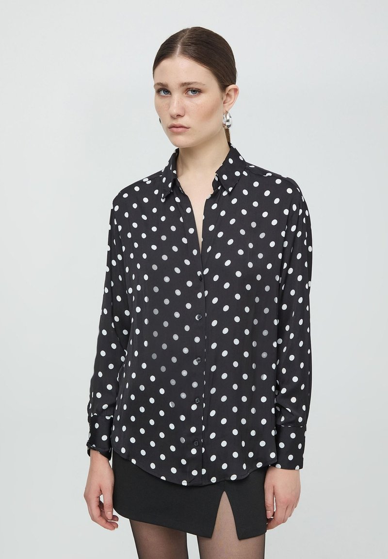 Camicia nera con bottoni, con pois bianchi, maniche lunghe, colletto e orlo leggermente curvo. Tessuto morbido e leggero.