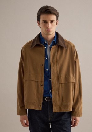 Jonge man met een bruin jack met een donkere kraag over een blauw overhemd met knopen en een spijkerbroek, staand tegen een effen beige achtergrond.