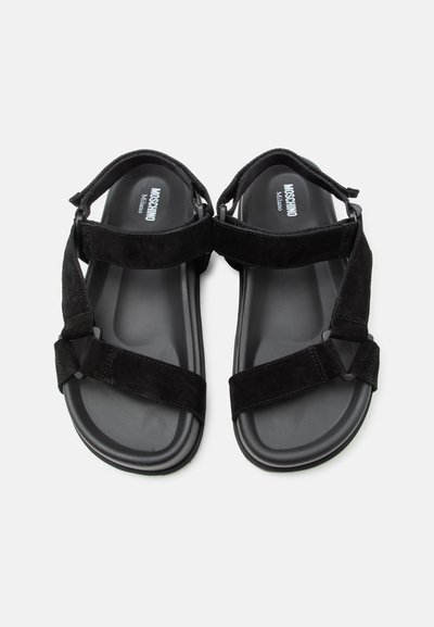 Μαύρα σανδάλια Moschino Milano με ρυθμιζόμενους ιμάντες βέλκρο και επενδυμένα πέλματα, απεικονίζονται από πάνω σε λευκό φόντο.