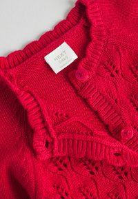 Rode gebreide babycardigan met een geschulpte kraag, decoratieve gaten en twee roze knopen. Label zegt "NEXT baby."