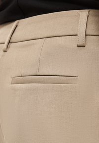 Κοντινή λήψη μπεζ υφάσματος που δείχνει μία πίσω τσέπη και θηλιές για ζώνη σε ραμμένο παντελόνι, φορεμένο με μαύρο τοπ.