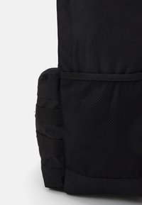 Canto de uma mochila preta com um bolso em malha texturizada, tecido reforçado e alças ajustáveis para fixar os itens. Design compacto e utilitário.