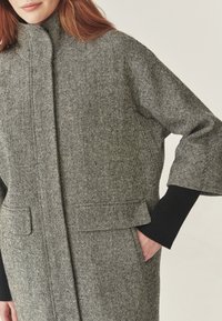 Cappotto in misto lana grigio con una texture a motivo herringbone, collo alto e tasche frontali. Presenta polsini a coste neri sulle maniche.