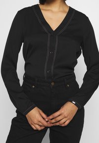 Blusa nera con maniche lunghe e scollatura a V, caratterizzata da cuciture a contrasto e chiusura con bottoni, abbinata a pantaloni neri a vita alta.
