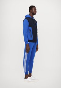 Tuta sportivo blu e nero con felpa con zip, tasche laterali e scarpe da ginnastica bianche con tre strisce lungo le gambe dei pantaloni.
