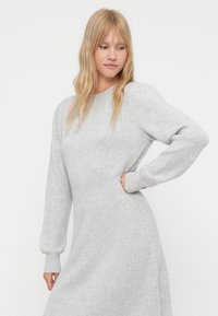 Donna con capelli biondi che indossa un vestito a maglia grigio chiaro con maniche lunghe, vita aderente e gonna texturizzata, in posa con una mano sulla vita.