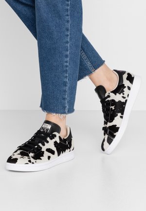 Person iført Adidas-sneakers med sort og hvidt ko-print kombineret med blå jeans med frynset kant, stående på et hvidt gulv.