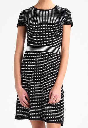 Femme portant une robe noire à manches courtes avec des rayures horizontales blanches en pointillés et une bande rayée blanche à la taille.