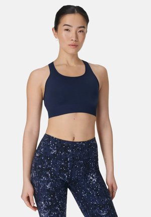 Vrouw die een marineblauwe sportbeha en marineblauwe, hoog getailleerde, patroonleggings draagt, staand met een neutrale uitdrukking tegen een effen achtergrond.