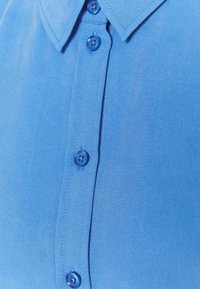 Jasnoniebieska koszula z detalami, w tym zapięciem na guziki, niebieskimi guzikami i klasycznym kołnierzykiem. Tkanina charakteryzuje się gładką teksturą.
