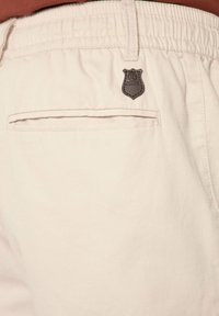 Shorts beige clair avec une taille élastique, dotés d'une poche cousue et d'un badge en cuir foncé sur la zone de la poche.