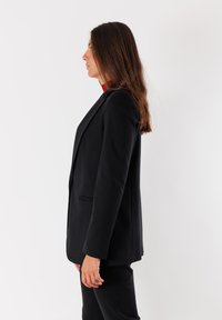 Donna con capelli castani che indossa un blazer nero e pantaloni, in piedi di profilo su uno sfondo bianco.