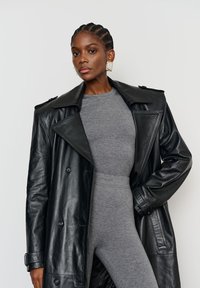 Fekete bőr trench coat nagy hajtókákkal és gombokkal, egy szürke, testhezálló overál fölé rétegzett. A modellen nagy négyzet alakú fülbevalók vannak.