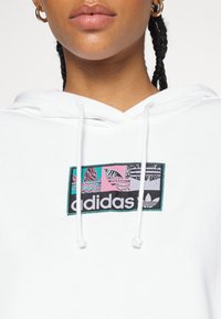 Lähikuva henkilöstä, joka on pukeutunut valkoiseen huppariin, jossa on värikäs Adidas-logo ja valkoiset narut, näyttäen huulet ja letitetyt hiukset.