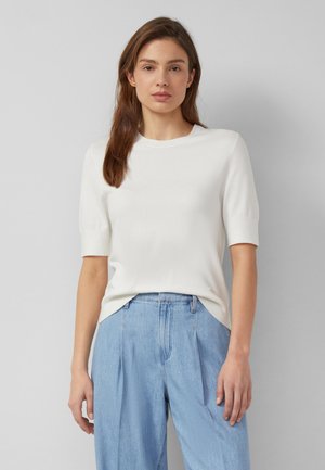 Kobieta o prostych brązowych włosach, ubrana w biały top z krótkim rękawem i jasnoniebieskie, szerokie spodnie z wysokim stanem, stojąca na szarym tle.