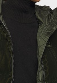 Lehká prošívaná olivově zelená bunda se zipem, přes fitted černý žebrovaný rolák. Hladká textilie a moderní design.