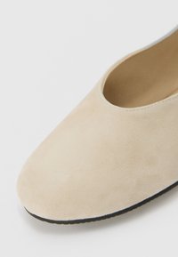 Beige Wildleder Slipper mit runder Zehenpartie und niedrigem Ausschnitt. Weiche Textur und schwarze Gummisohle mit gewelltem Rand.