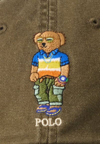 Figura de urso bordada em tecido verde-oliva. O urso veste uma camisa azul, laranja e branca, calças cargo, óculos de sol e acessórios. "POLO" embaixo.