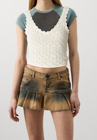 Bílý háčkovaný crop top s vlnitými okraji, navrstvený přes světle modrošedé tričko s krátkým rukávem, doplněný o vybledlé džínsové sukně s sklady.