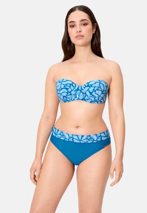 Mujer con un bikini azul sin tirantes con patrones de hojas blancas en la parte superior y la cintura, de pie contra un fondo claro y liso.