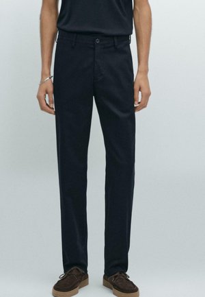 Pantalon bleu marine en tissu lisse, présentant une coupe droite, des poches avant et une fermeture à bouton. Porté avec des chaussures en suède marron.