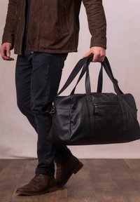 Black leather duffel bag with dual handles and a zippered pocket, carried by a person in dark jeans and brown shoes. Smooth texture and structured design.