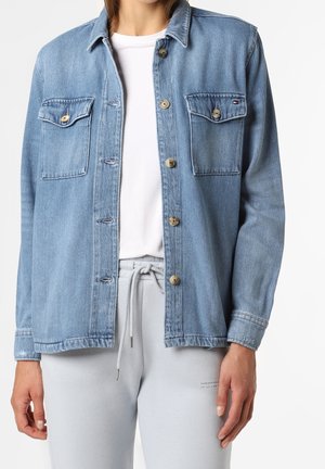 Person wearing a light blue denim jacket over a white shirt and light gray drawstring pants, standing against a plain background.