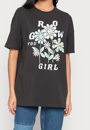 Czarny t-shirt z pastelowymi uśmiechającymi się kwiatami i tekstem "Grow you girl", noszony przez osobę w niebieskich dżinsach.