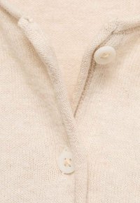 Beige gestrickte Strickjacke mit weichem Material, V-Ausschnitt und zwei weißen Knöpfen am Hals. Kompakter und leichter Stoff.