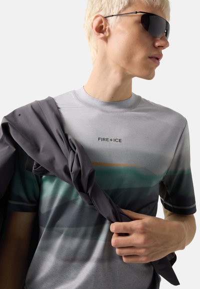 Junge Person trägt schwarze Sonnenbrille, ein grau-grünes Verlaufs-Shirt mit dem Schriftzug „FIRE + ICE“ und hält eine dunkle Jacke über einer Schulter.