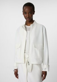 White jacket with a mandarin collar, zip closure, two front pockets, and ribbed texture. Worn over a white ribbed top.