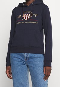Granatowa bluza z kapturem z miękkiego materiału, z przednią kieszenią, kapturem ze sznurkiem oraz haftowanym logo w kolorach czerwonym i złotym. Luźny krój.