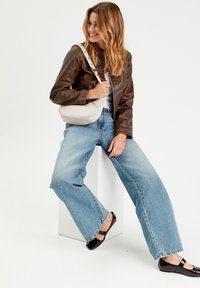 Lichtgrijze schoudertas, bruine leren jas, witte top, losse blauwe jeans, zwarte ballerina's. Model poseert zittend op een witte sokkel.