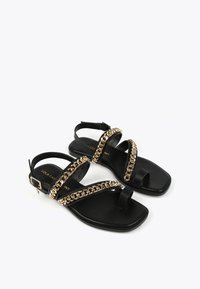 Mustat litteät sandaalit, joissa on ristikkäiset hihnat ja kultaketjua korostava yksityiskohta, soljetaan nippusoljen avulla, ja niissä on teksturoitu pohja. Neliömäinen kärki.